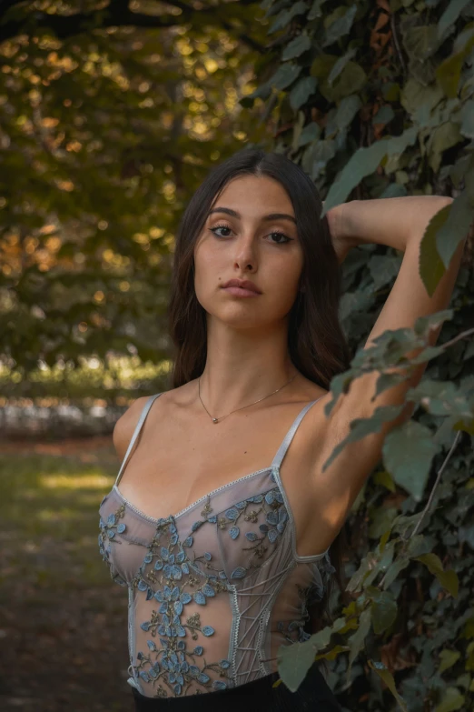 a woman in an open - top shirt leans against a tree