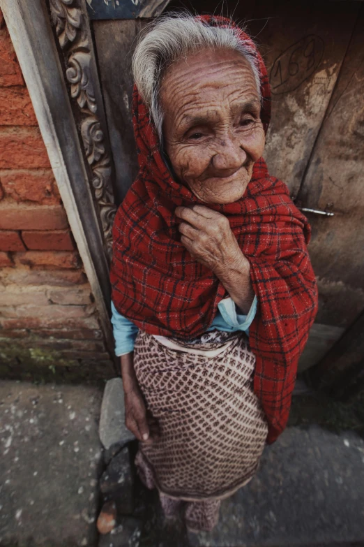 an old woman with no makeup wearing a red scarf, with a cigarette in her mouth and a wooden frame holding up soing