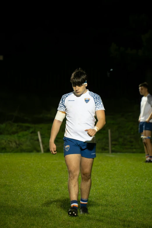 a young man in white and blue soccer uniform is in grass at night with a ball