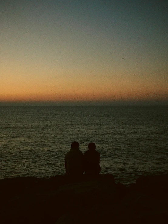 two people sitting on the rocks, while the sun sets