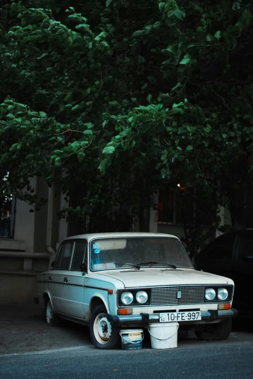 a white car with a dented headlight on parked under a tree