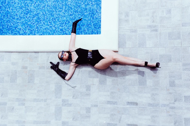 a woman in black swimsuit laying on a tile floor next to a swimming pool