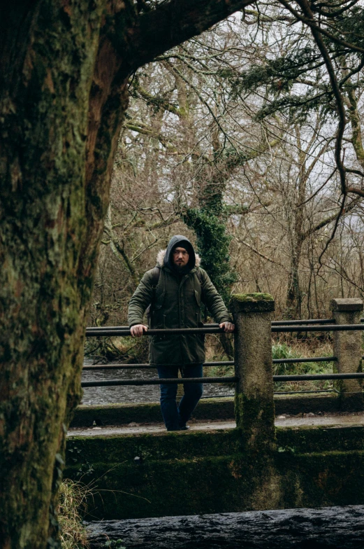 man with hooded jacket standing on fence surrounded by trees