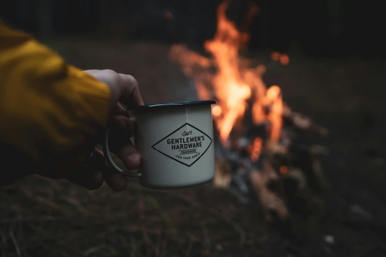 someone is holding a coffee mug next to a campfire