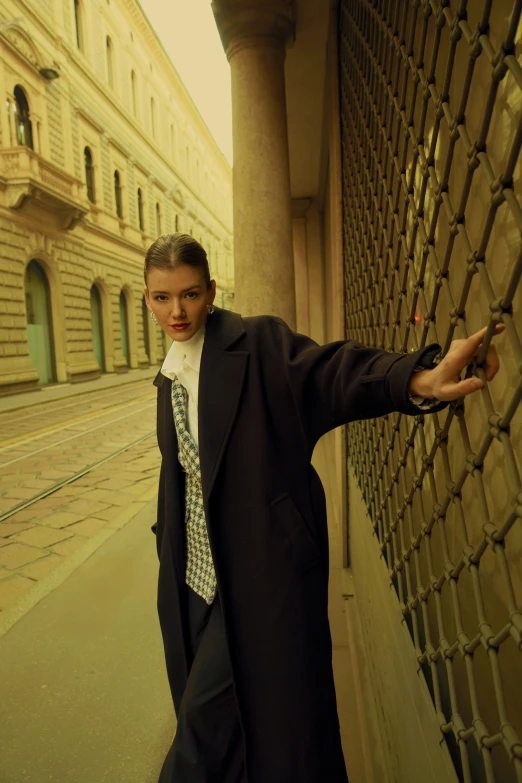 a young man wearing an overcoat leaning against a fence