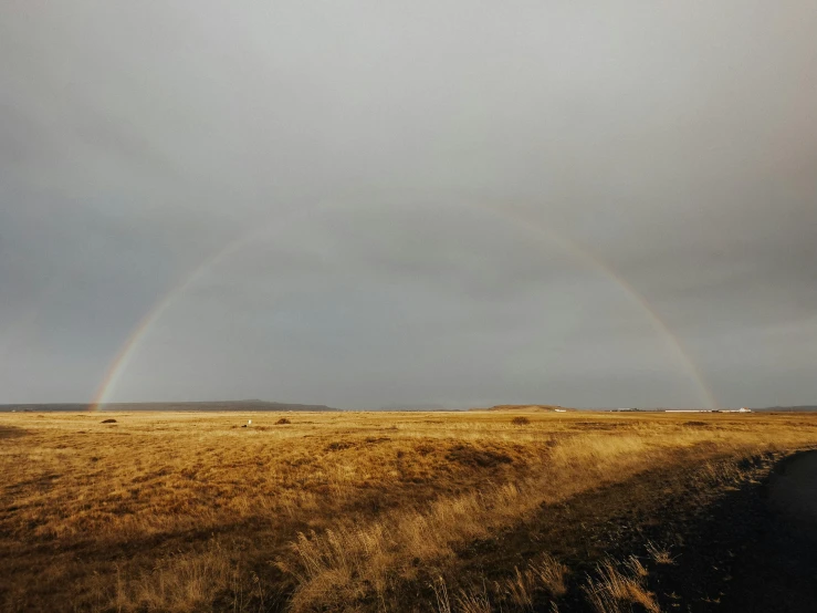 two rainbows shine in the dark gray sky as a dirt road sits at the end