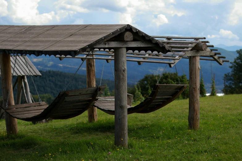 two wooden swings in a grassy area with mountains and clouds behind