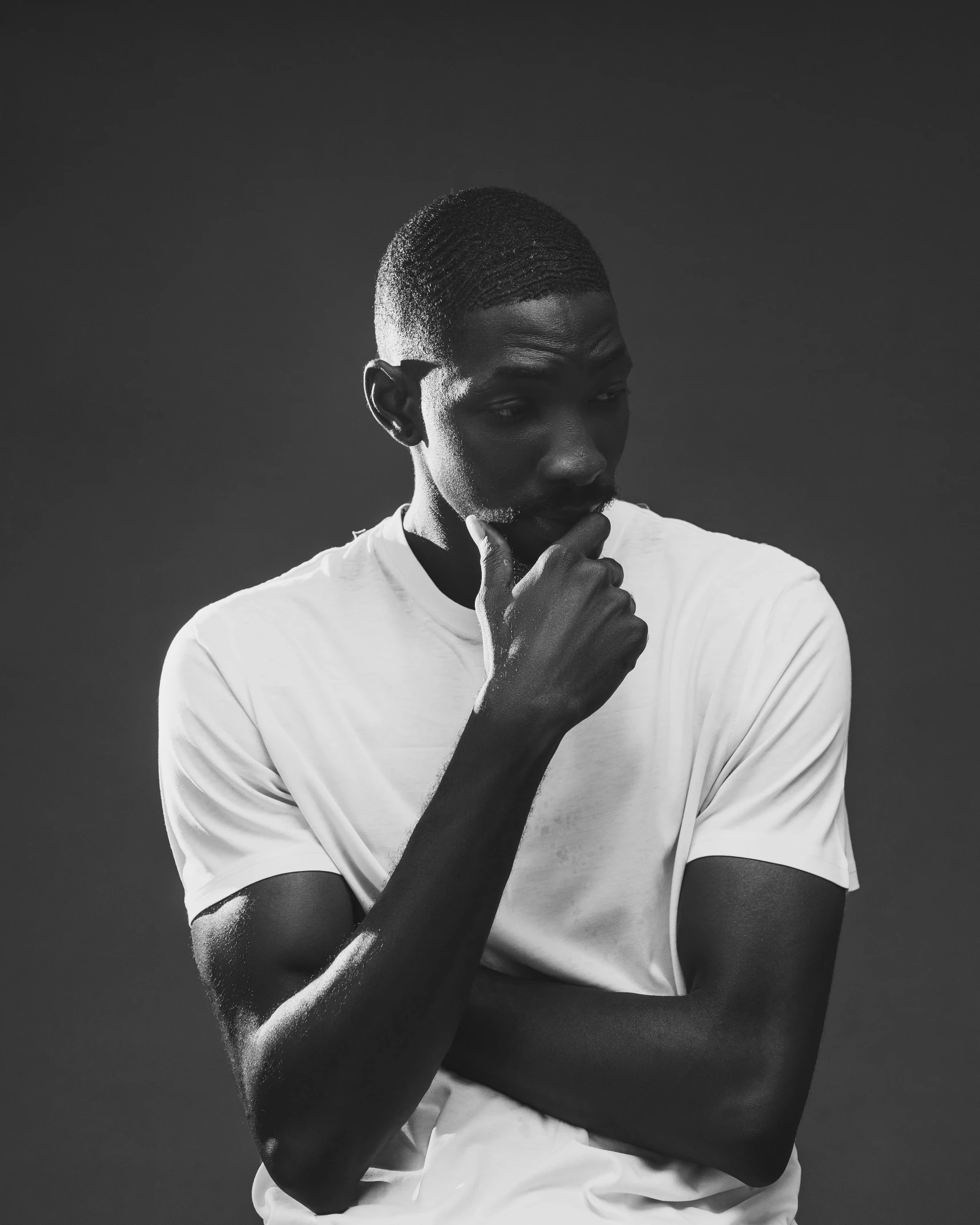 a black man wearing a white shirt stands looking down and covers his face