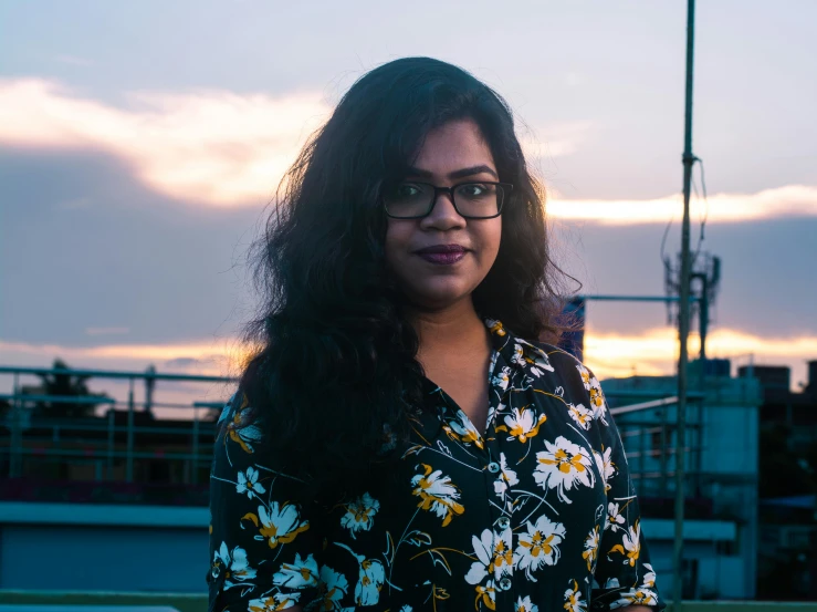 a woman with long dark hair and glasses is standing outdoors at sunset