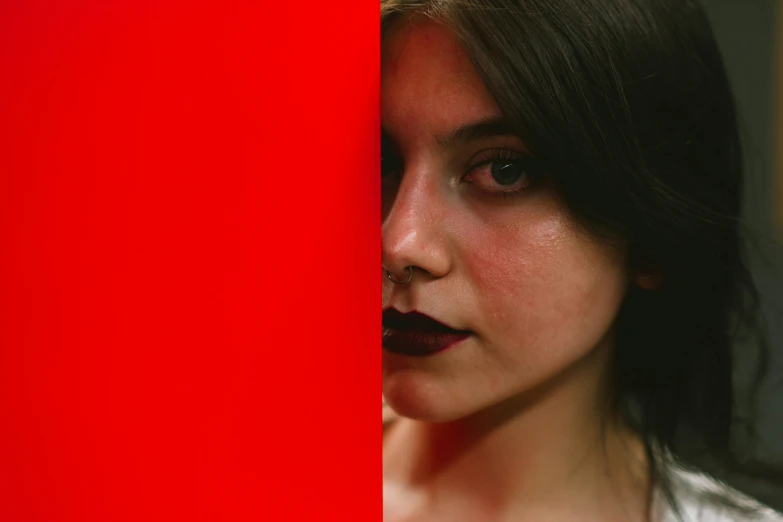 a woman with dark makeup peering out from behind a red wall