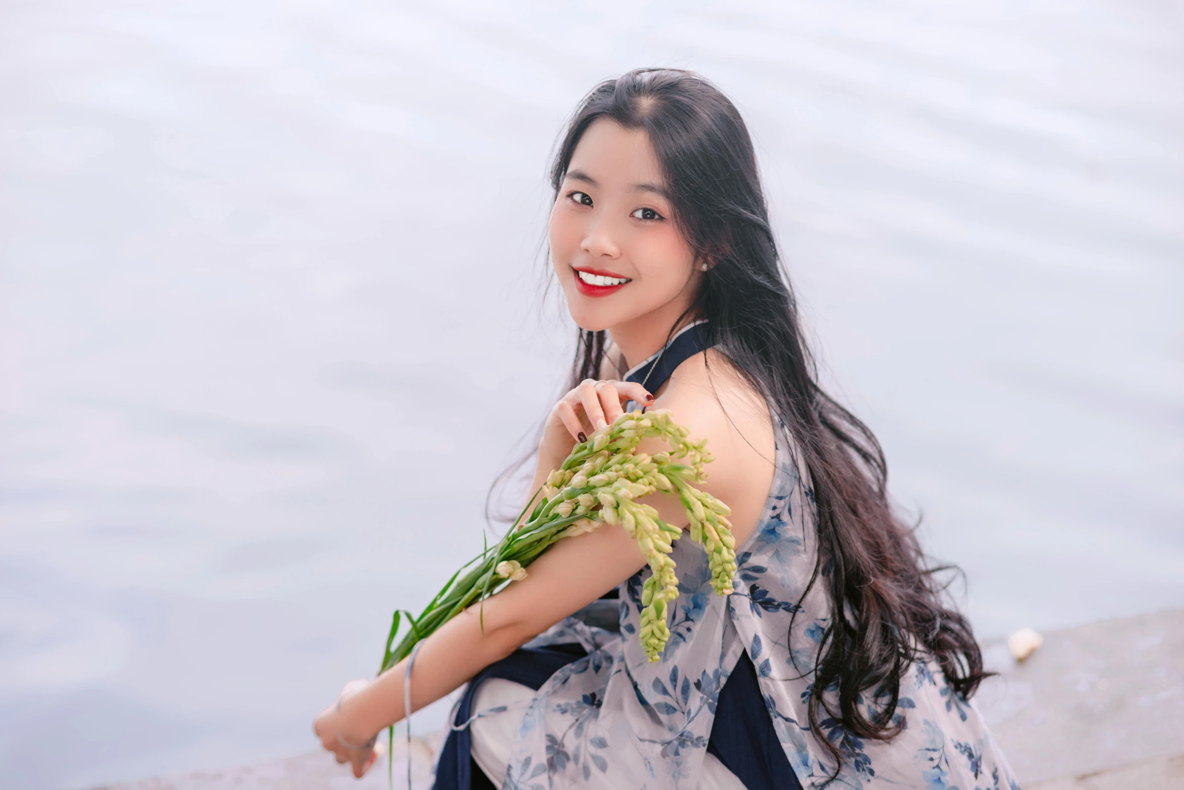 a woman smiles while holding flowers by a body of water