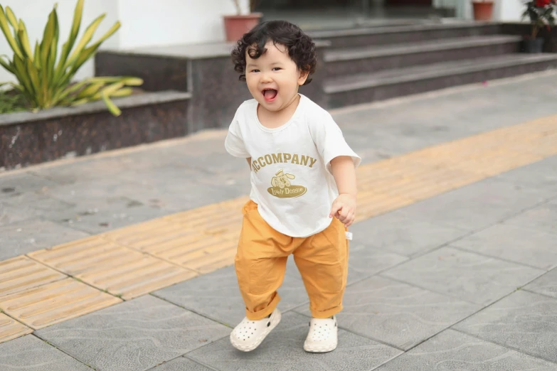 an adorable little boy in brown pants and a white t - shirt standing with one foot on his hip