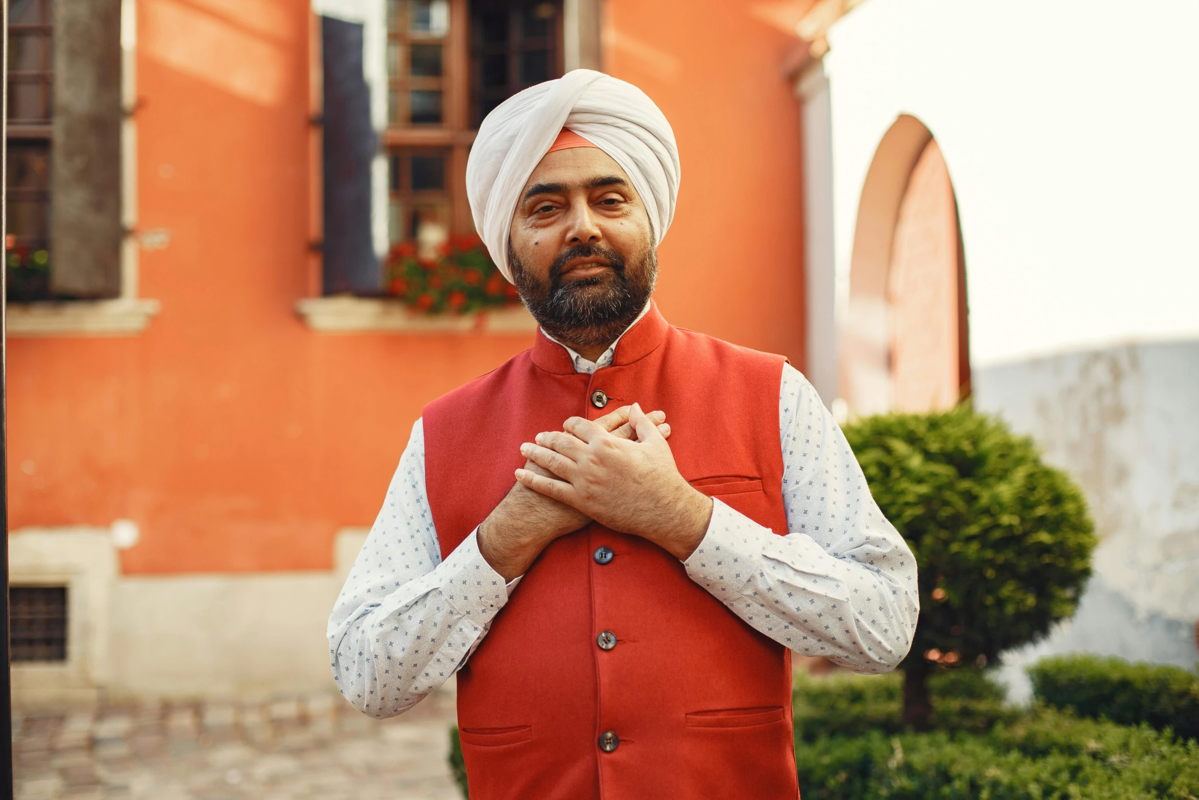 a man dressed in a turban with the hands folded, standing in front of an orange building