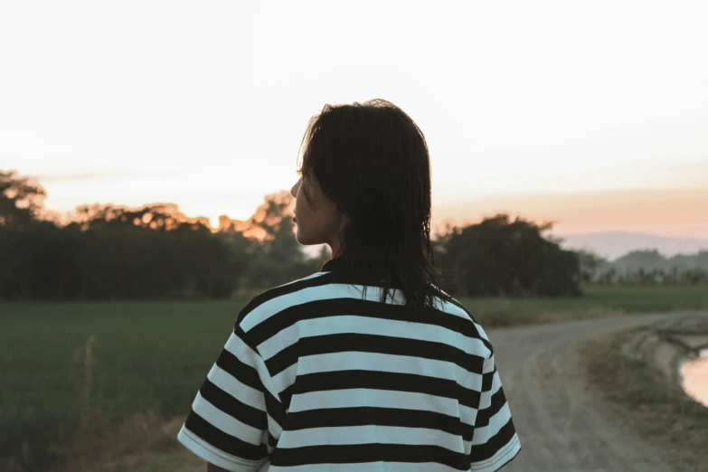 a girl in a striped shirt is standing on a dirt road and looking at a sunset