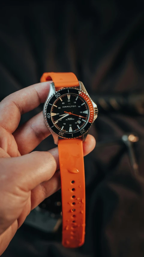 a hand is holding an orange watch in it's right hand