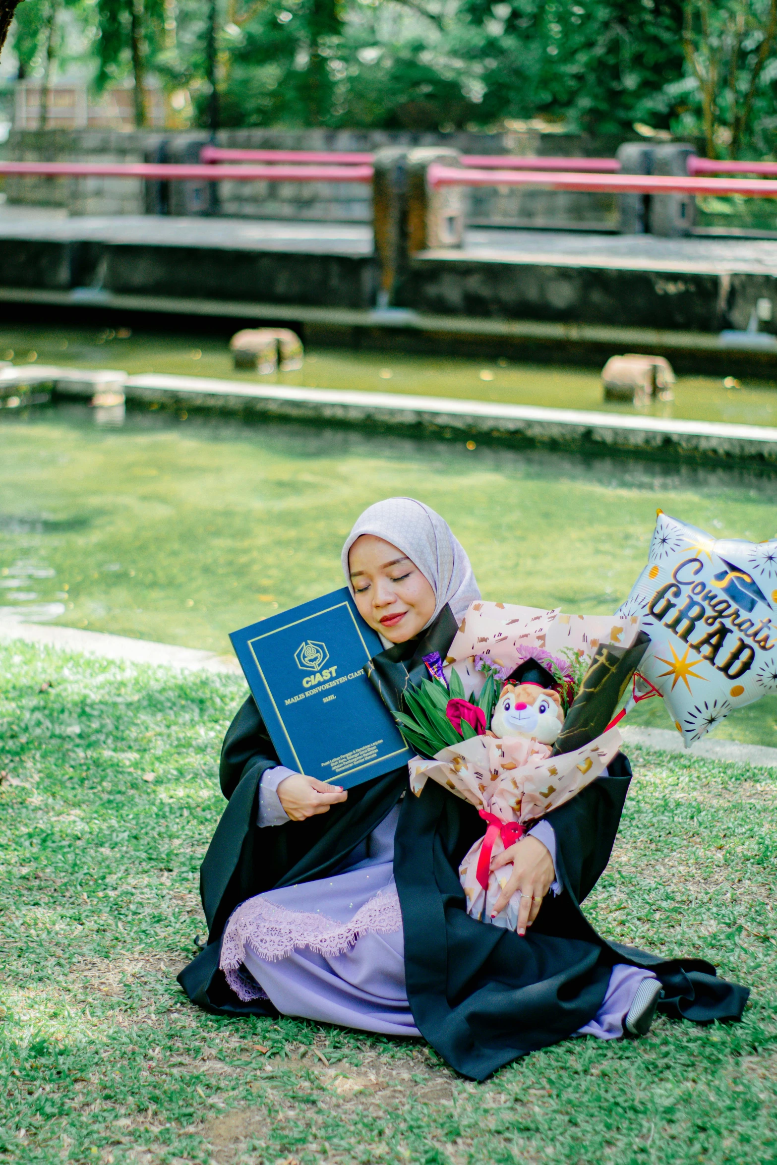 a woman with a graduation cap sits on the grass and holds an award