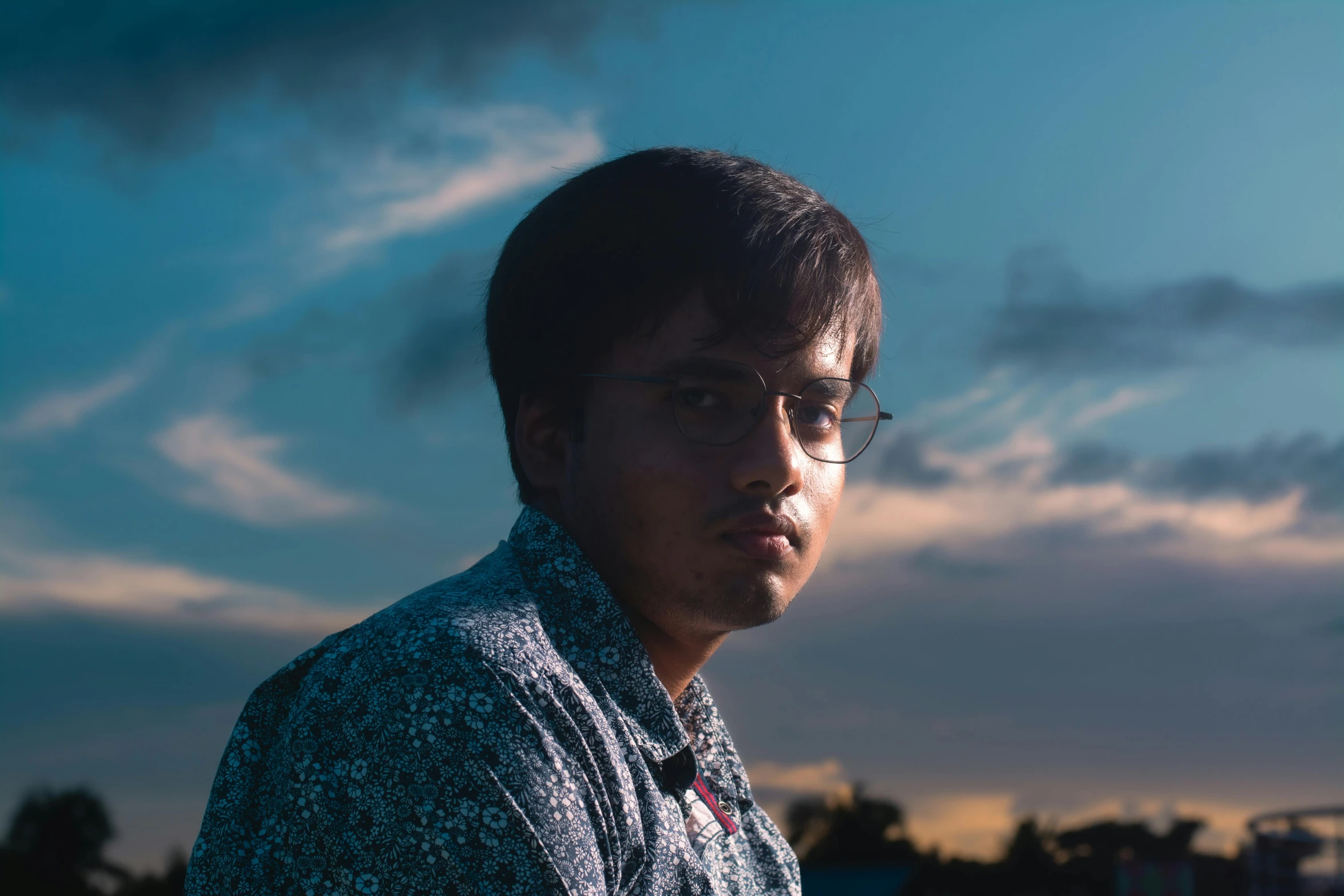 a man wearing glasses, standing in front of some clouds