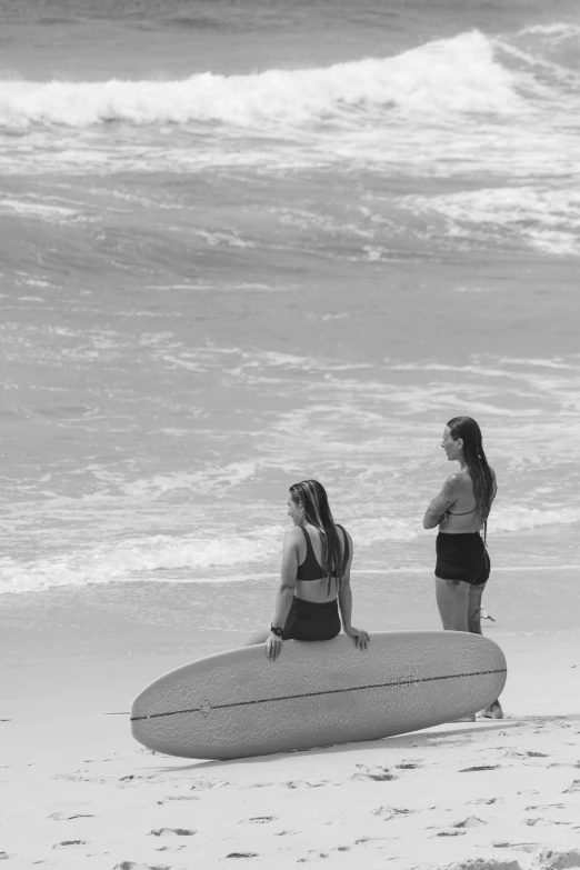 two women standing on the beach with a surfboard