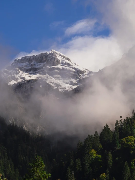 a mountain with some snow on top and clouds blowing in the air