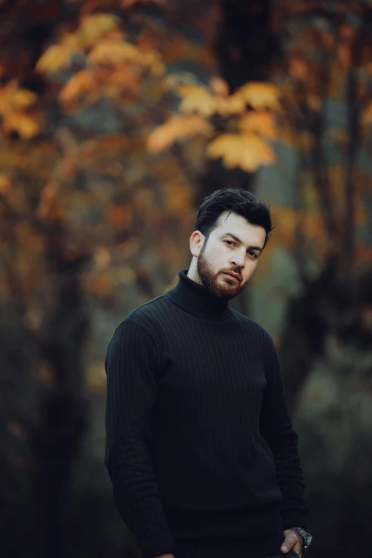 a young man wearing black stands by a tree with fall foliage