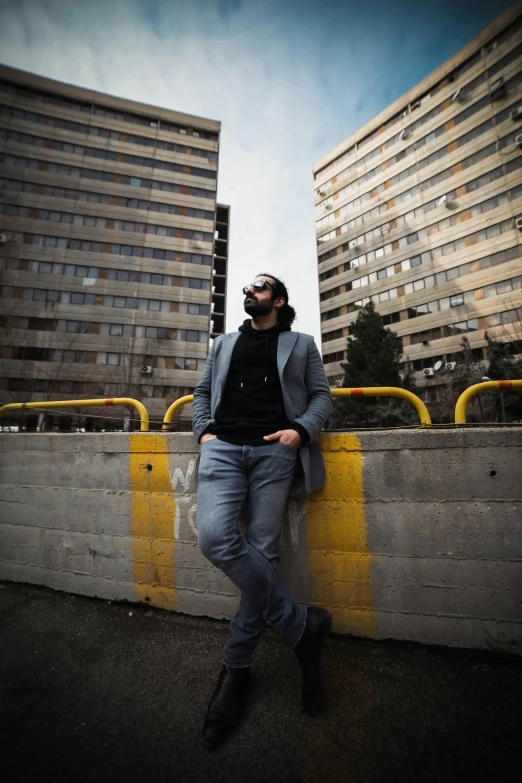 a bearded man leans against the concrete barrier in front of two building