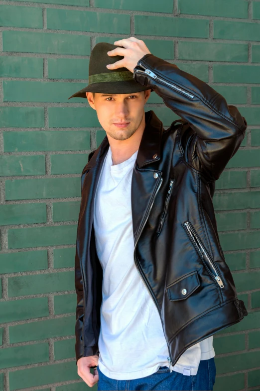 a man in a leather jacket and hat looks back with hand on his head