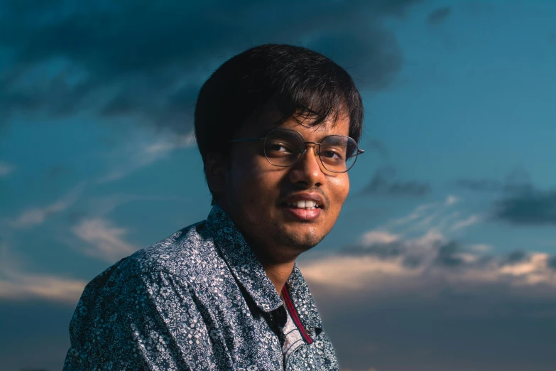 a man in glasses standing in the dirt under a cloudy sky