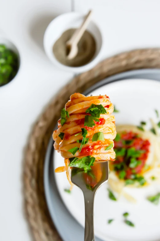 fork full of pasta on white plate with small bowls of sauce and pepper sprinkled with green leafy foliage