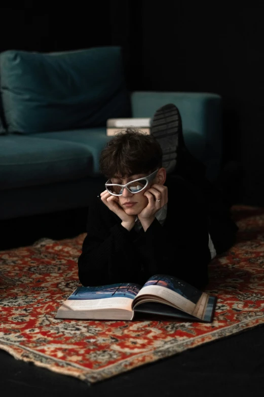 a person with glasses sitting on a rug and reading a book