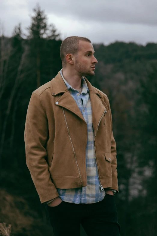 a man in a jacket and plaid shirt is standing near a forest