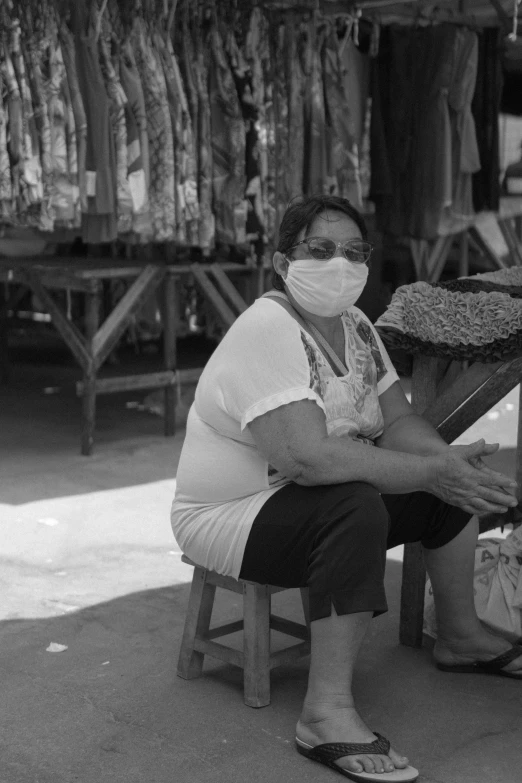a woman with a face mask sits on a stool at a market