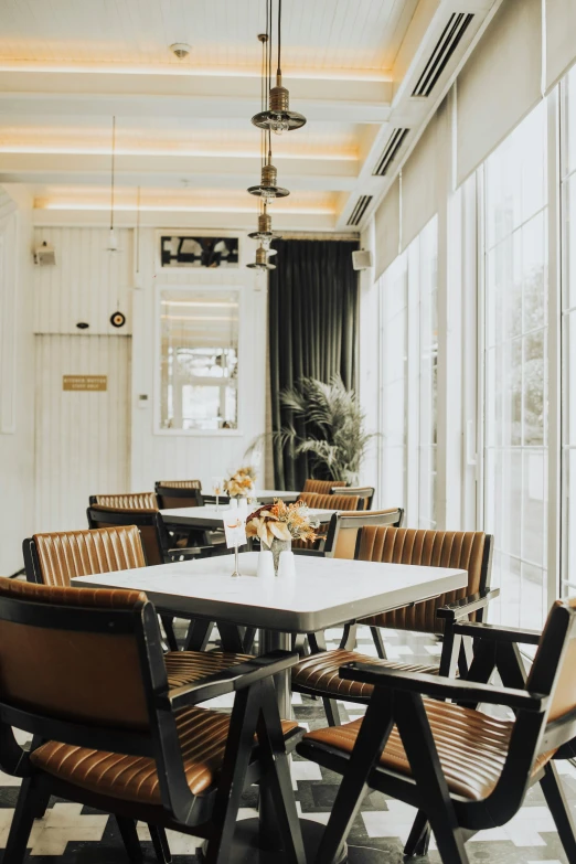 the interior dining area of a modern el with white walls, wooden beams, and high ceiling, has large windows, a table and tan leather chairs