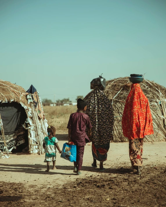 two women carrying bags are walking with a child in front of huts