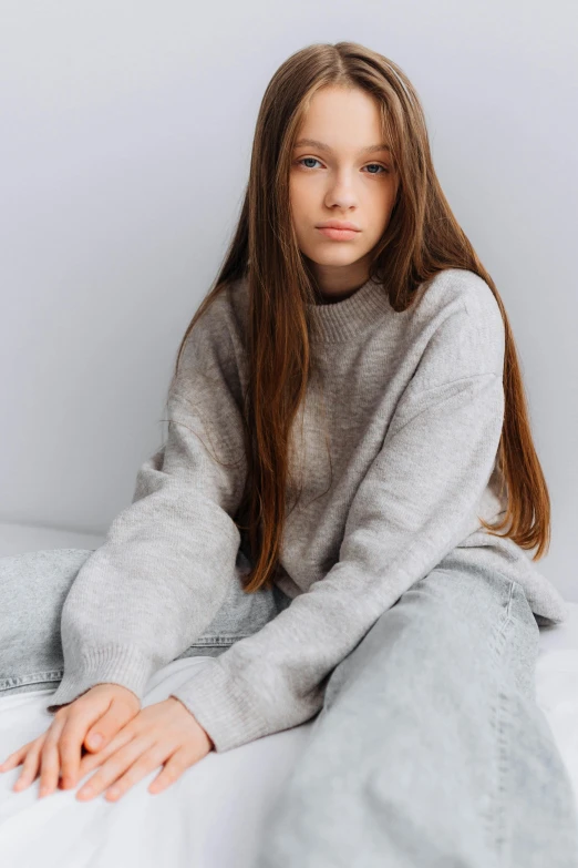 woman with long, straight hair wearing grey sweater sitting on a bed