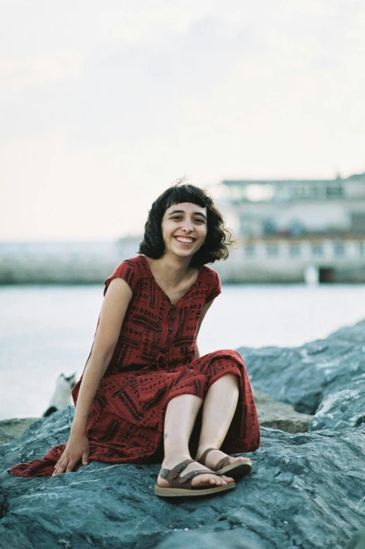 a woman in a red dress sitting on the rock by the water