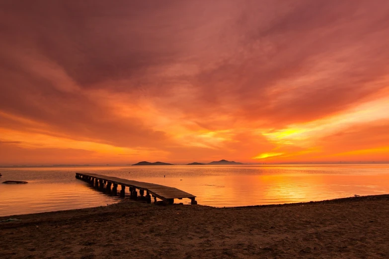 a pier sitting on top of a sandy beach next to a body of water, a picture, by Mathias Kollros, shutterstock, sunset red and orange, greece, very very wide shot, autumn sunset