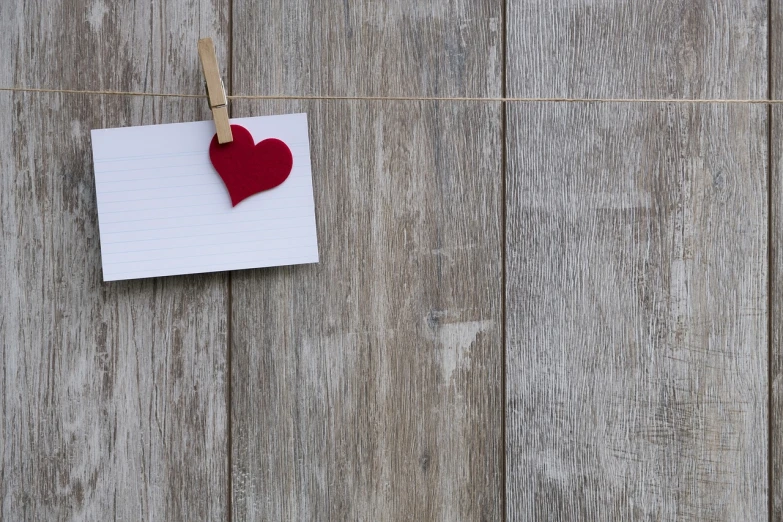 a piece of paper with a red heart hanging on a clothes line, a picture, trending on pixabay, minimalism, wooden background, hidden message, still life photo of a backdrop, looking across the shoulder