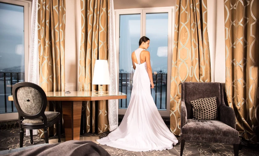 a woman in a wedding dress standing in front of a window, inspired by Adrienn Henczné Deák, hotel room, tanned ameera al taweel, fancy drapery on furniture, white silky outfit