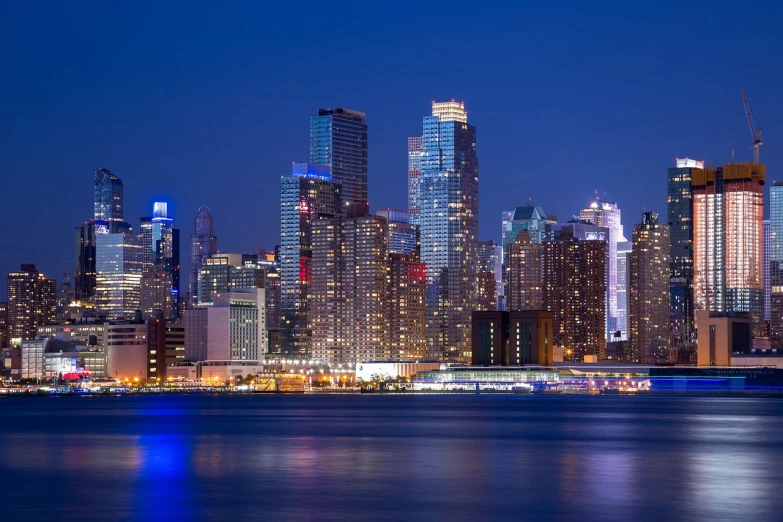 a large body of water with a city in the background, a photo, by William Berra, shutterstock, new york city at night, blue hour lighting, with shiny glass buildings, modern high sharpness photo