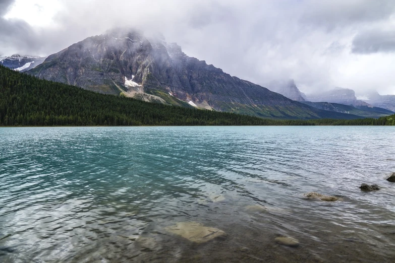 a body of water with a mountain in the background, a picture, by Raymond Normand, shutterstock, overcast lake, puddles of turquoise water, low ultrawide shot, smokey water scenery