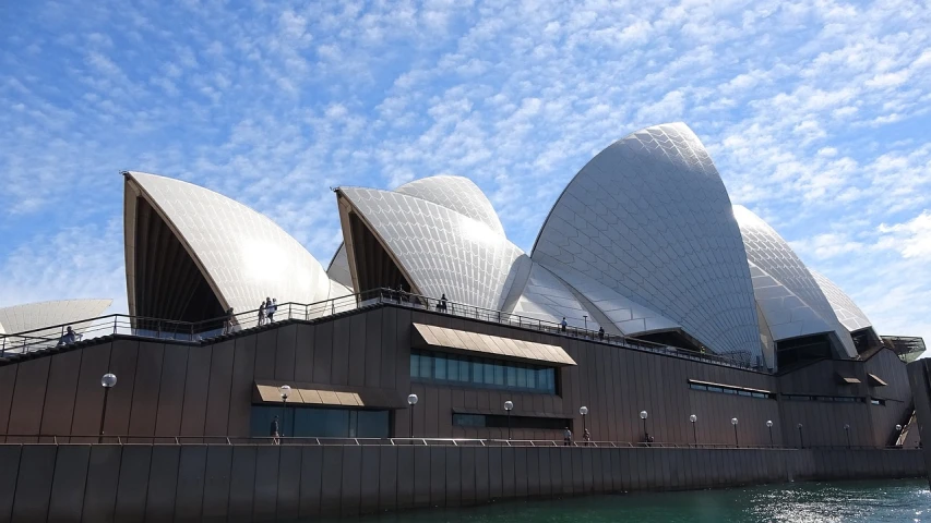 a large building sitting next to a body of water, a picture, inspired by Sydney Carline, flickr, modernism, singing at a opera house, blue skies, huge support buttresses, photo taken in 2018