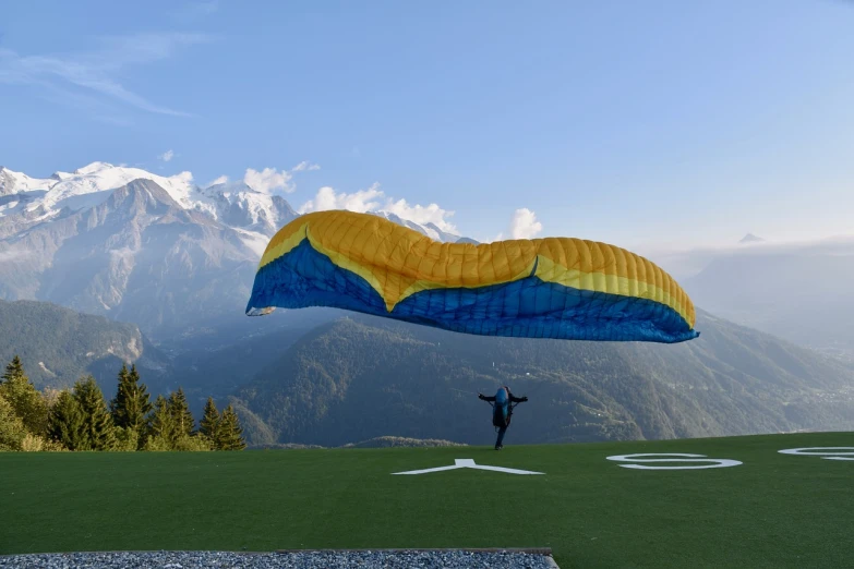 a man standing on top of a lush green field, a picture, figuration libre, parachutes, the alps are in the background, 2 0 2 2 photo, aerodynamic imposing