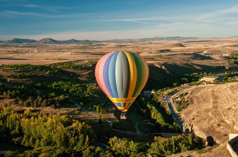 a colorful hot air balloon flying over a valley, a picture, by Pedro Pedraja, shutterstock, madrid. extreme long shot, drone shot, historical setting, summer evening