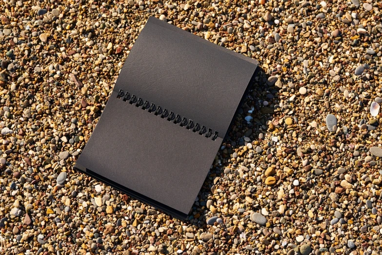 a black notebook sitting on top of a gravel covered ground, by Jan Rustem, highly detailed product photo, on the sand, neoprene, high quality paper