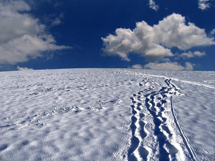 a person riding skis down a snow covered slope, a photo, flickr, “puffy cloudscape, footprints, blue sky with beautiful clouds, whorl. clouds