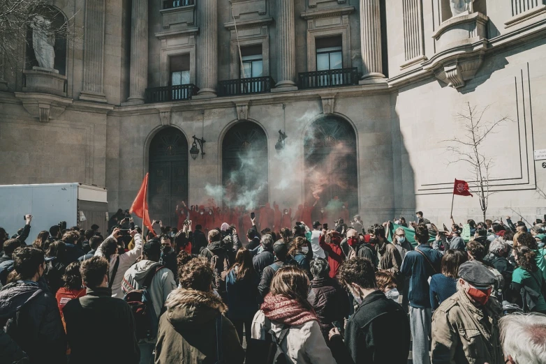 a crowd of people standing in front of a building, by Luis Molinari, pexels, figuration libre, red smoke, beware the ides of march, academism, in the middle of the day