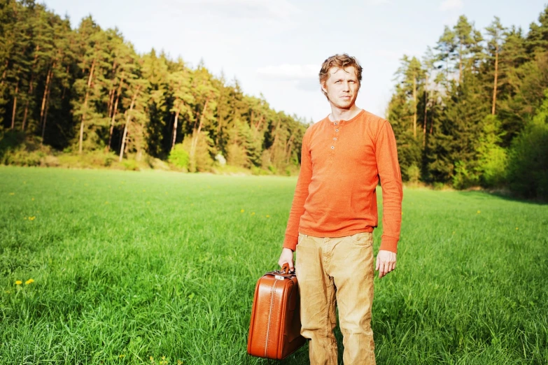 a man standing in a field holding two suitcases, a stock photo, by Kurt Roesch, shutterstock, wearing an orange t shirt, analogue photo, near forest, portrait of young man