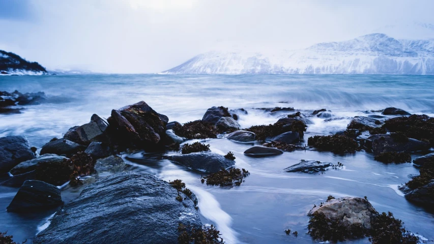 a body of water with rocks and a mountain in the background, a picture, inspired by Johan Christian Dahl, pexels, wintery scene, sea spray, 4 k smooth, shore