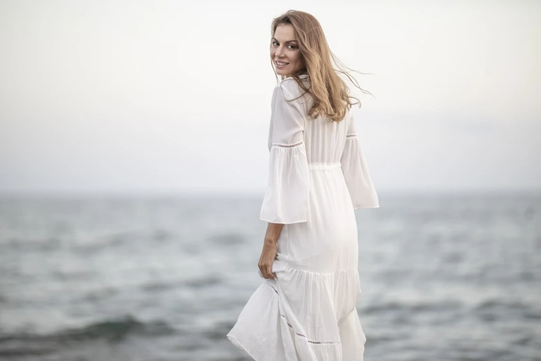 a woman standing on top of a beach next to the ocean, inspired by Antonín Chittussi, shutterstock, romanticism, white! dress, long tunic, three quarter profile, close up portrait shot