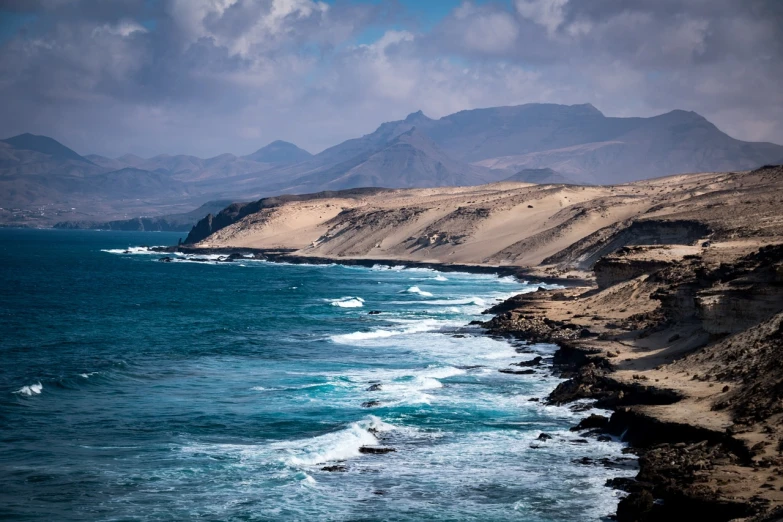 a large body of water with mountains in the background, a tilt shift photo, by Alexander Robertson, shutterstock, les nabis, windy beach, black volcano afar, spain, hawaii beach